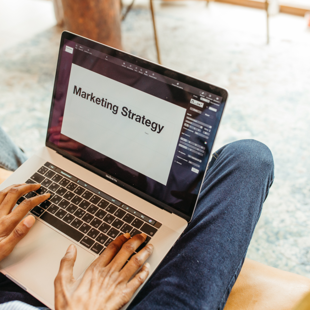 Someone sitting with a laptop on their lap, the laptop is the focus, and the words MARKETING STRATEGY are written across an otherwise blank page on-screen