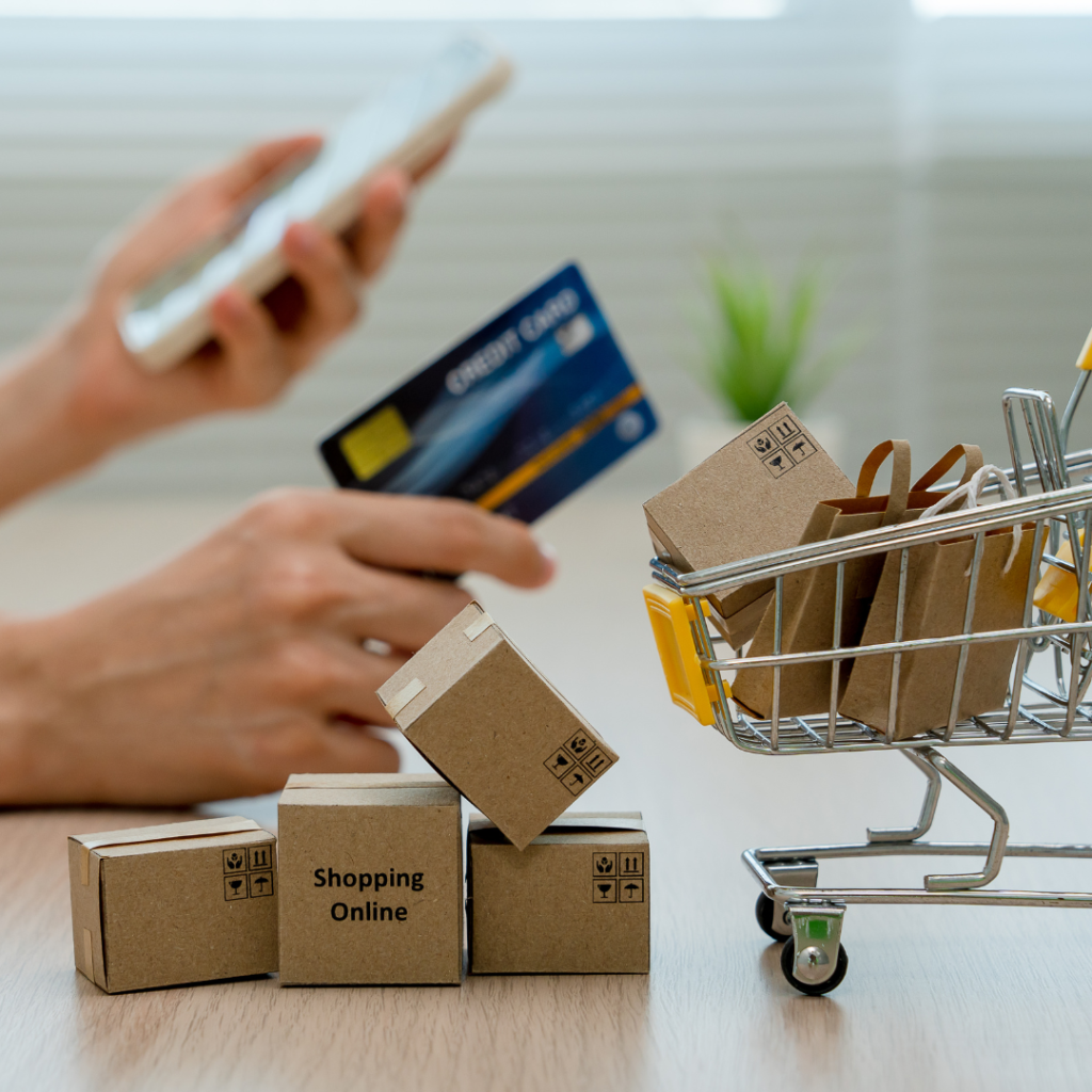 A tiny shopping cart with little cardboard boxes that say "shopping online", and in the background, a woman holding a credit card in one hand and her phone in the other. 