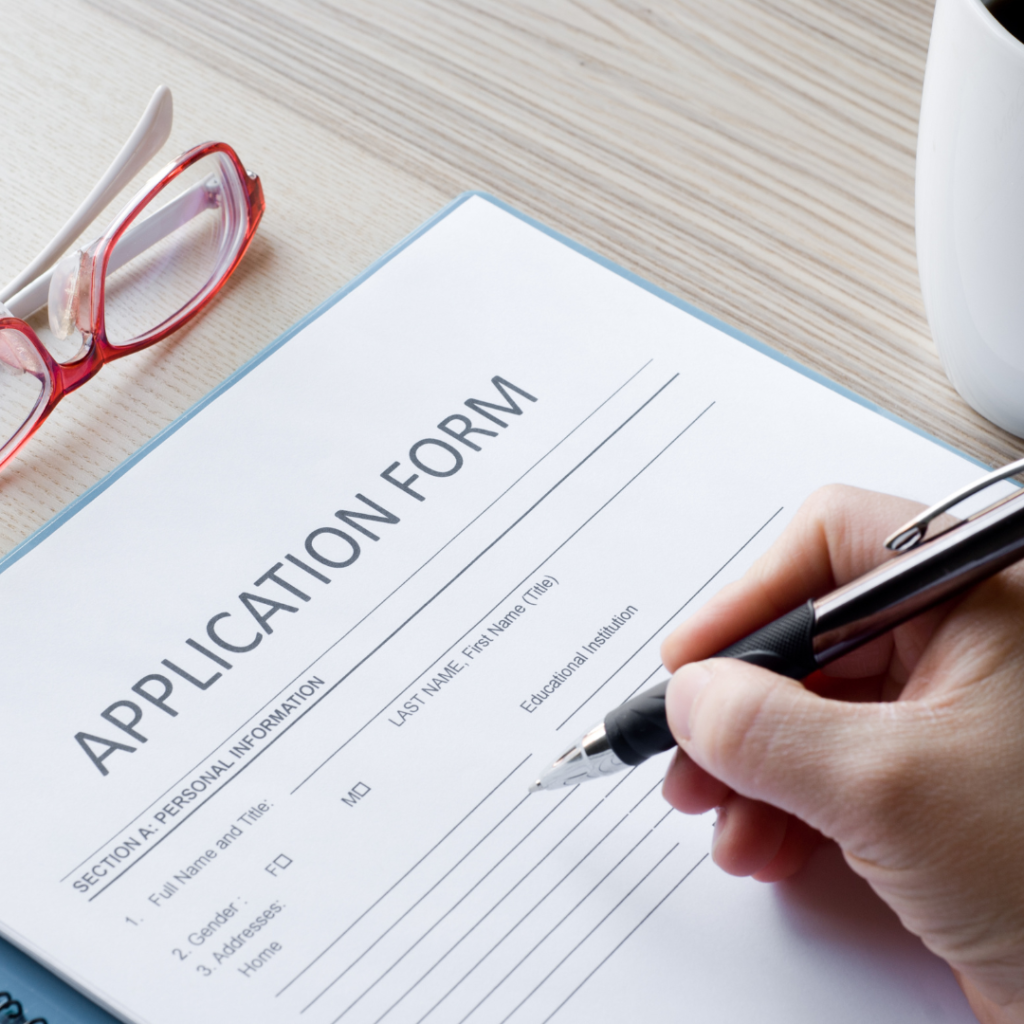 Application form, pen poised over page ready to fill it out, glasses folded on the table above the forms. 